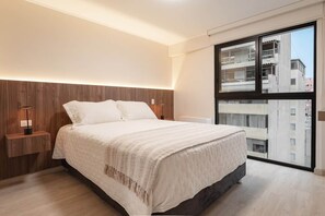 A modern bedroom with a large bed, bedside tables, a nightstand, and a large window with a view of buildings. - High-Floor 1BR · A/C, Pool & View · Miraflores (Miraflores)