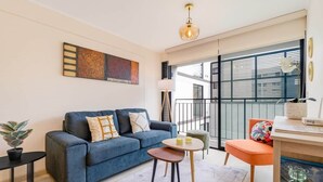 Un salon moderne avec un canapé bleu, une petite table ronde, une plante en pot et une vue sur l’extérieur grâce à de grandes fenêtres.