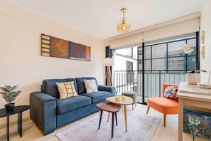 Ein modernes Wohnzimmer mit einem blauen Sofa, einem kleinen runden Tisch, einer Topfpflanze und einem Blick ins Freie durch große Fenster.