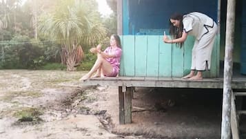 Una persona seduta su un portico con struttura in legno, un'altra appoggiata alla struttura, e un ambiente naturale con vegetazione.