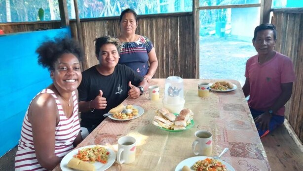 Un grupo de personas sentadas a una mesa con comida y bebidas, en una habitación con pared de madera y una ventana.
