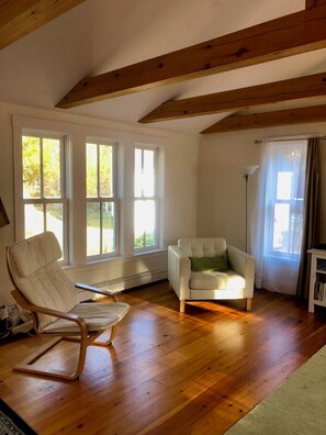 A room with wooden floors, a white armchair, a sofa, and large windows with curtains.