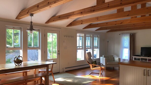 A spacious room with wooden beams, a dining table with benches, a sofa, a TV, and large windows offering a view of greenery.