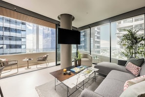 A modern living room with a large window, a flat-screen TV, a sofa, a coffee table, and a potted plant. - BlueMall Luxury Suites by Homeclub (Santo Domingo)