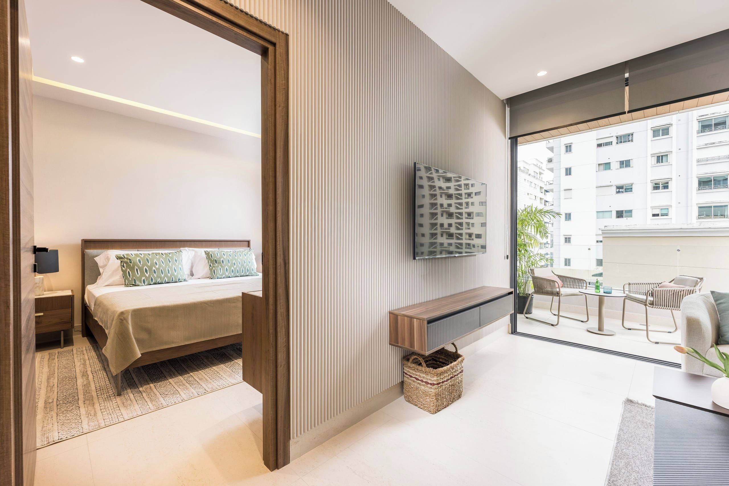 Une chambre d’hôtel moderne équipée d’un lit, d’un téléviseur fixé au mur, d’un balcon avec des sièges et offrant une vue sur le paysage urbain.