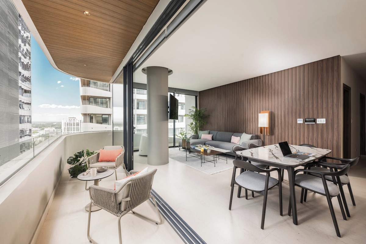 A modern living room with a sofa, a coffee table, and a dining table with chairs. There is a balcony with a view of the city.