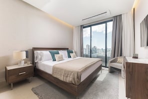 A modern hotel room with a large bed, a desk, a chair, and a view of the city. - BlueMall Luxury Suites by Homeclub (Santo Domingo)