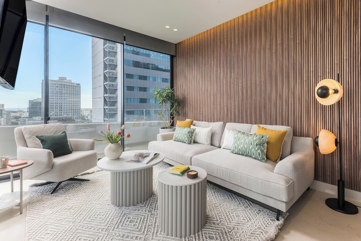 A modern living room with a sofa, two side tables, a coffee table, and a view of the cityscape through large windows.