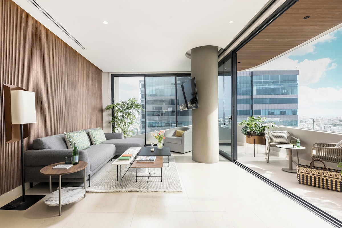A modern living room with a sofa, coffee table, and a view of a cityscape through large windows.