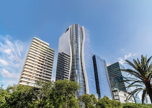 A modern high-rise building with a curved glass facade, surrounded by other tall buildings and lush green trees. - BlueMall Luxury Suites by Homeclub (Santo Domingo)
