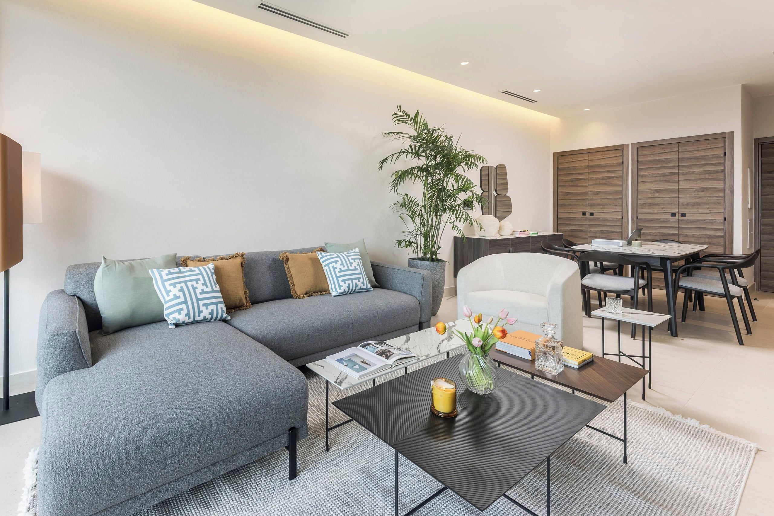 Un salon moderne avec un canapé d’angle gris, une table basse, un coin repas avec une table et des chaises, et une grande plante.