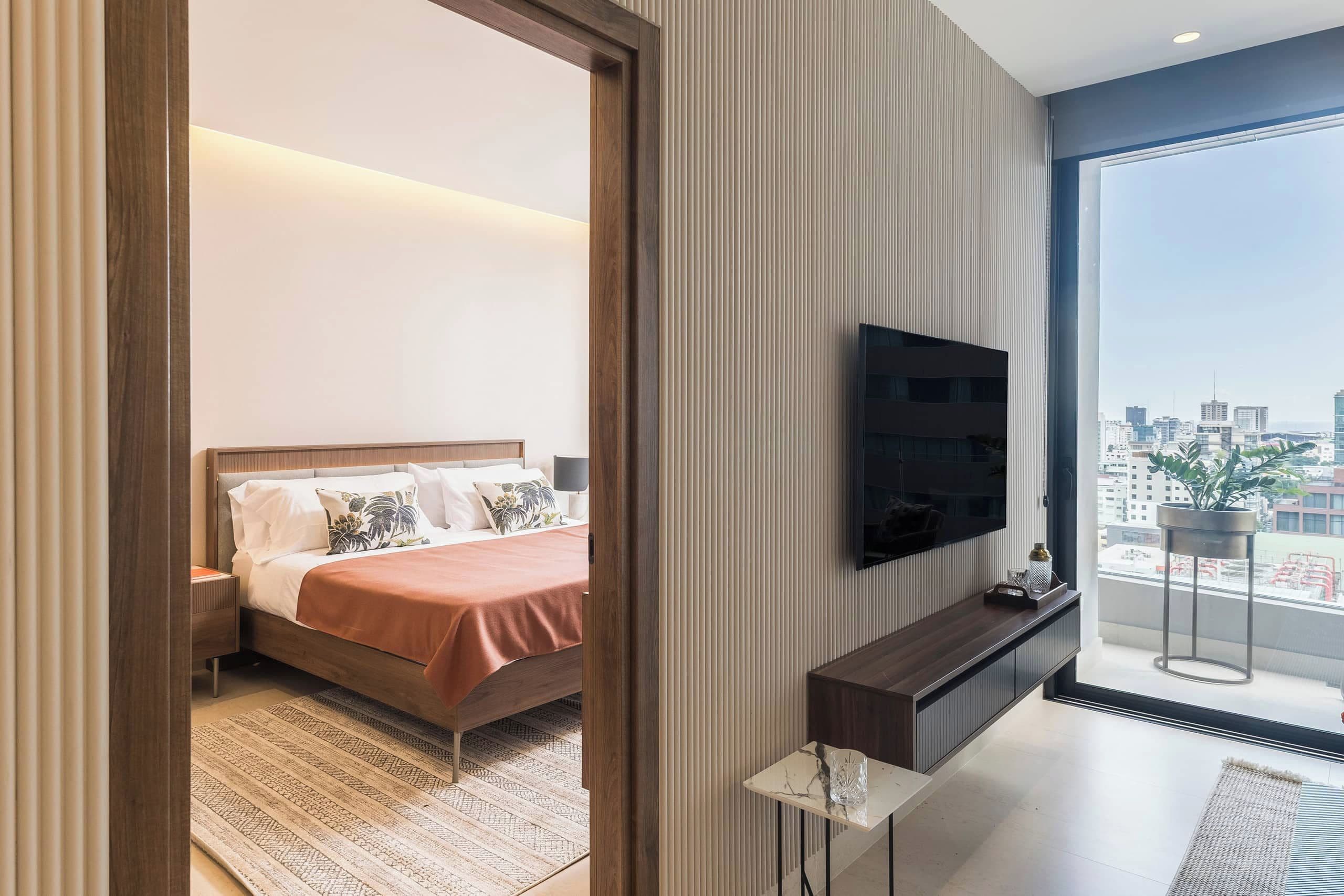 Une chambre d’hôtel moderne équipée d’un lit, d’une télévision, d’une petite table et d’une grande fenêtre donnant sur la ville.
