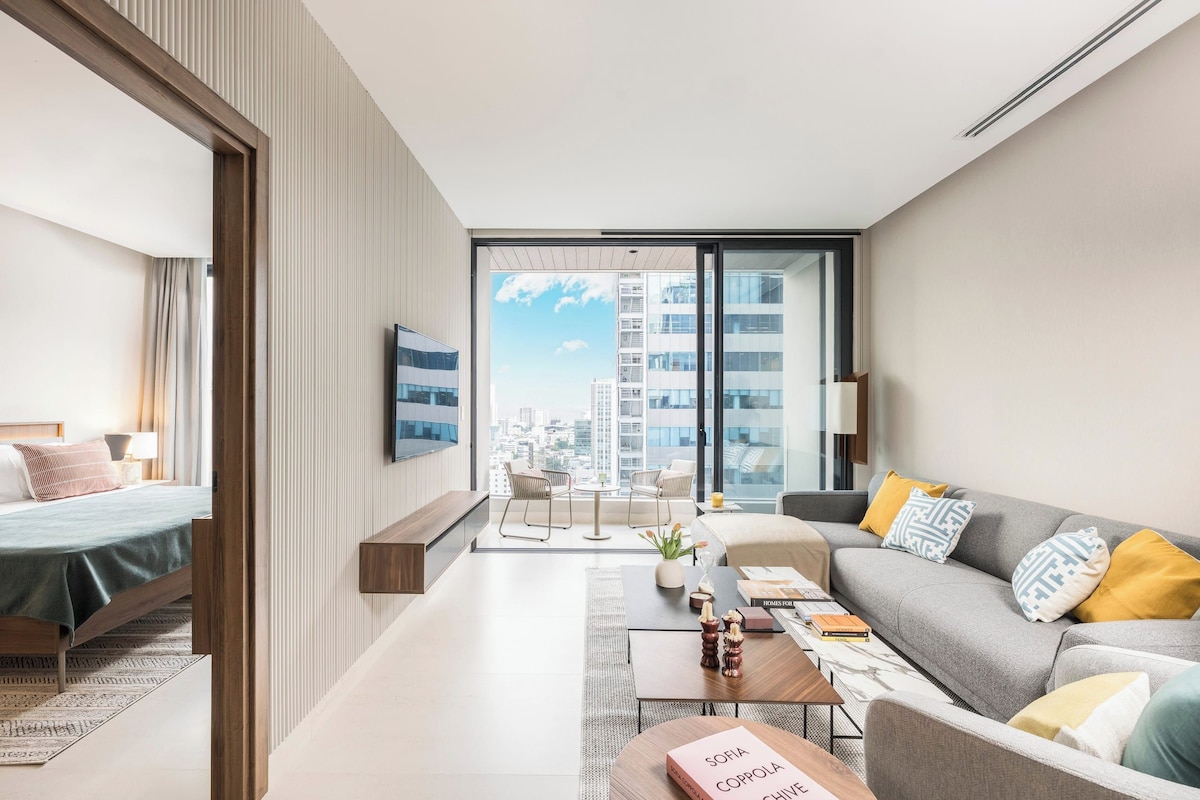 A modern hotel room with a bed, a sofa, a coffee table, and a view of the cityscape through large windows.