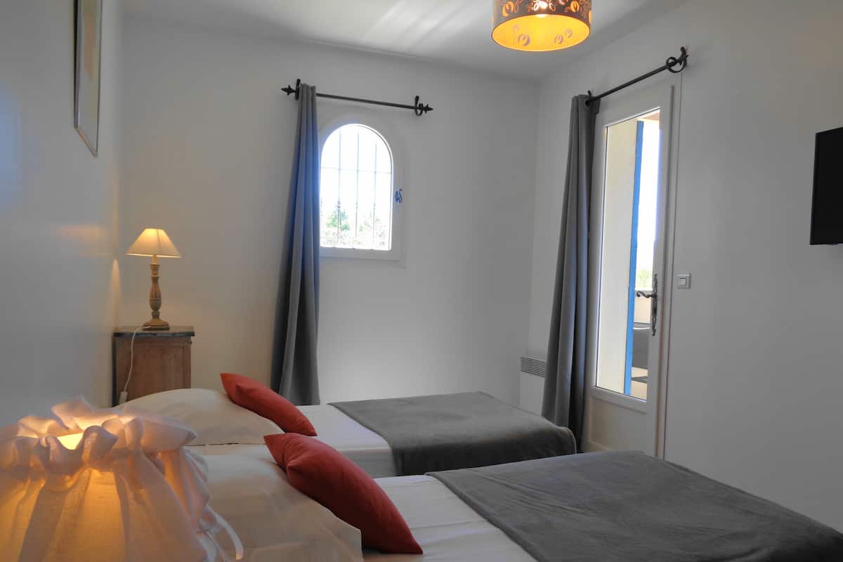A room with two beds, a window with curtains, a lamp, and a door leading outside.