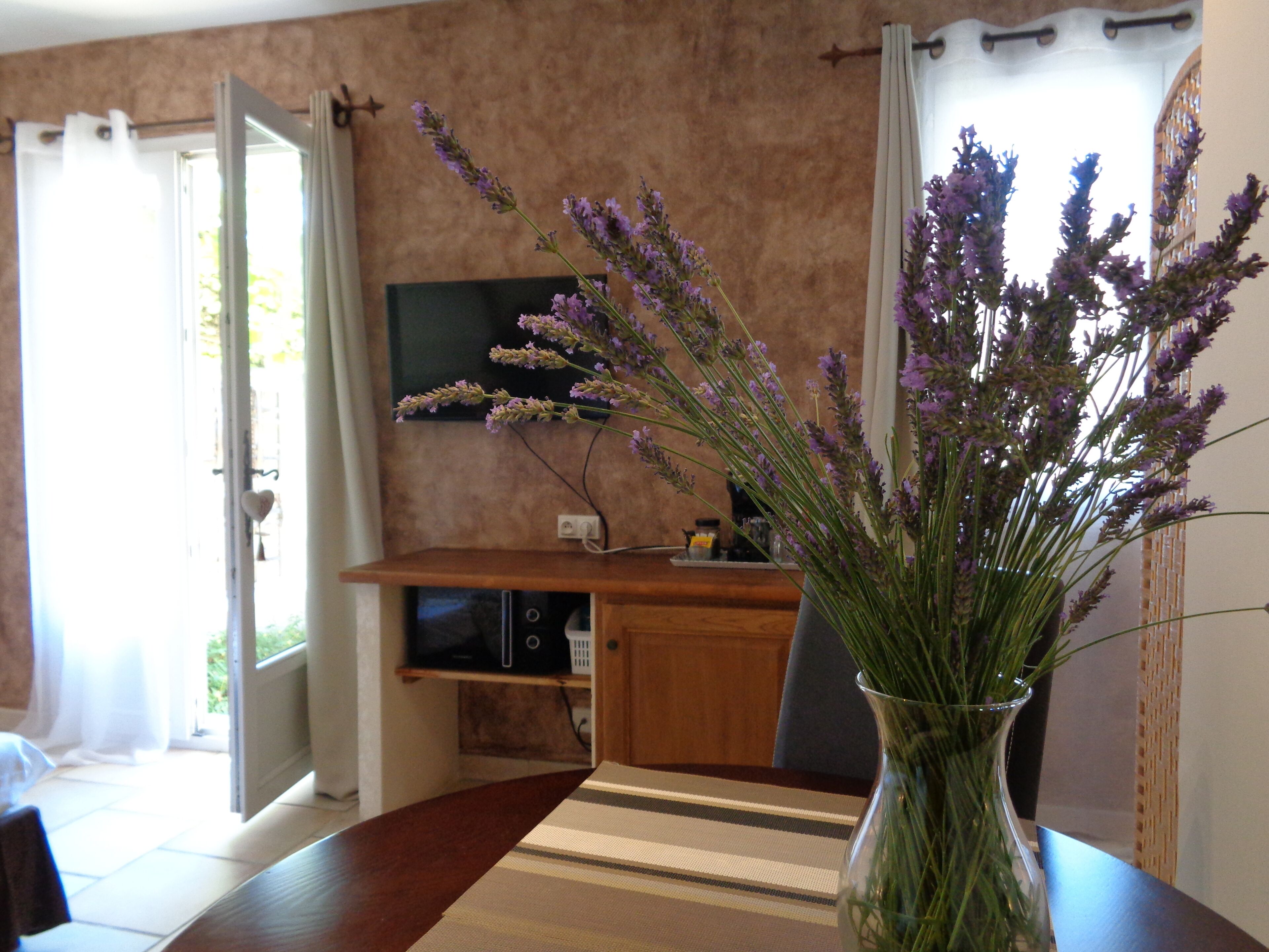 A dining area with a table, chairs, a vase with flowers, a television, and a kitchen area in the background.