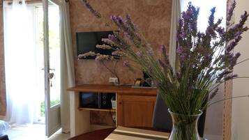 A dining area with a table, chairs, a vase with flowers, a television, and a kitchen area in the background.