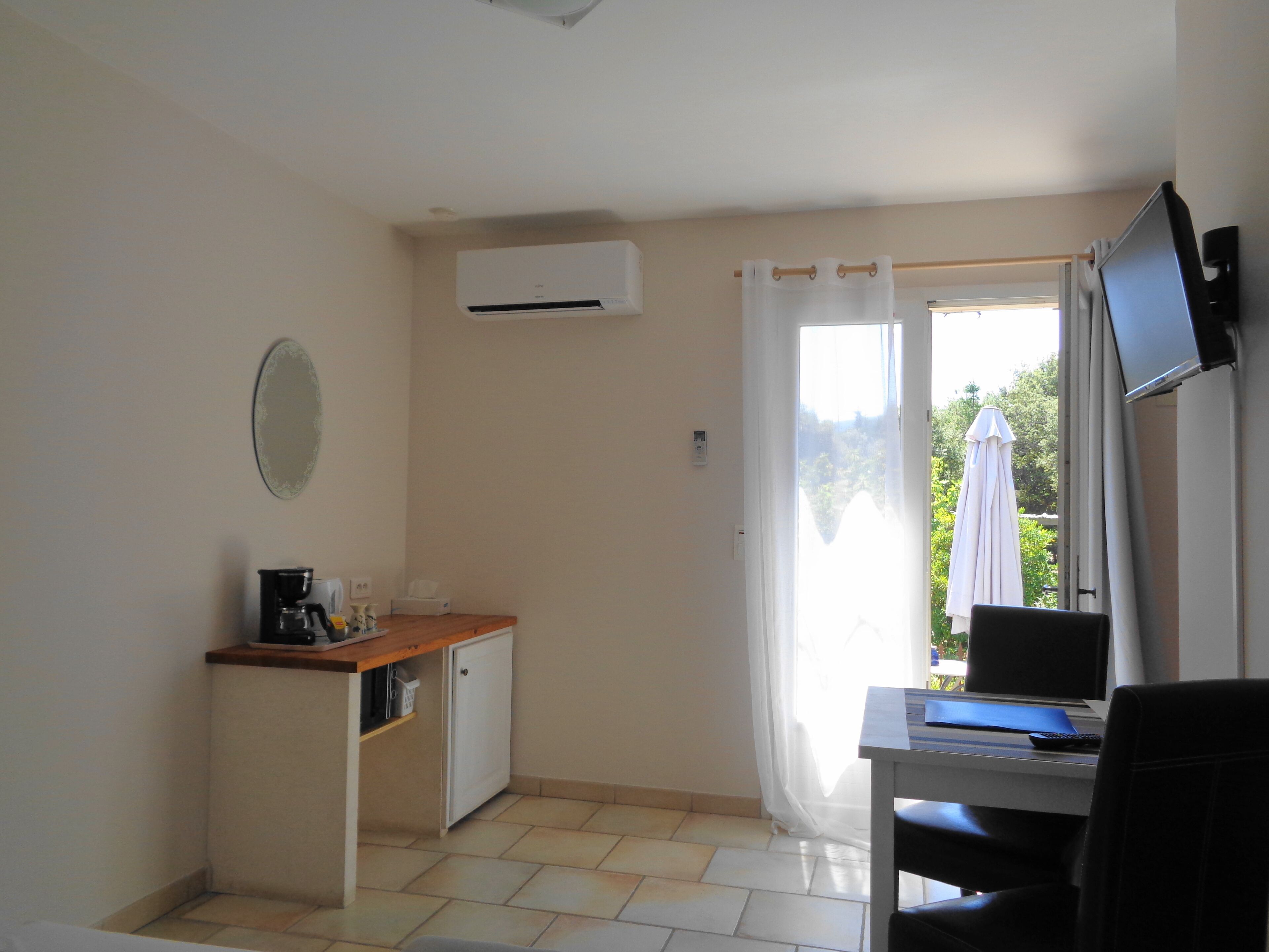 Een kamer met een bureau, een stoel, een televisie, een airconditioner, een klein kastje en een raam met gordijnen.