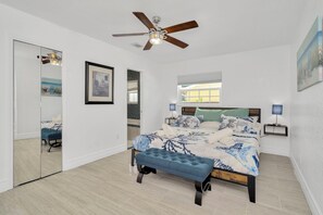 Ein Schlafzimmer mit einem Bett, einem Deckenventilator, einem Nachttisch mit Lampe, einem Spiegel und einem Fenster.