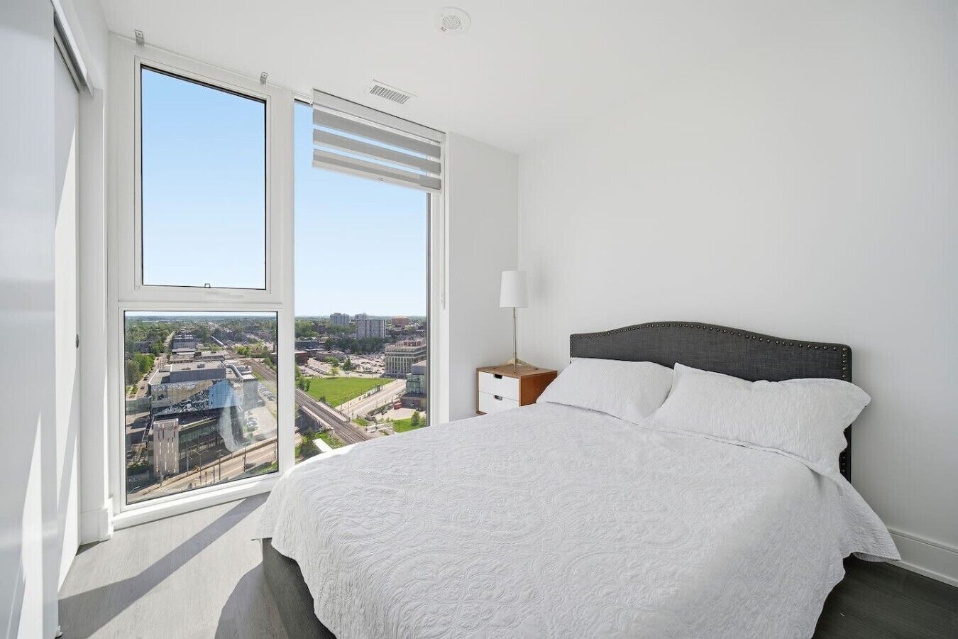 Un dormitorio con un ventanal grande, una cama con sábanas blancas, una mesita de noche con una lámpara y vistas a la ciudad.