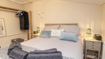 Una cama bien hecha con almohadas y toallas dobladas, una lámpara de noche, una mesita de noche con una cesta y un perchero en la pared.