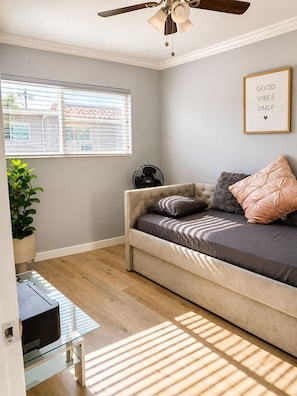 Ein Zimmer mit Deckenventilator, einem Fenster mit Jalousien, einer Couch mit Kissen, einer Topfpflanze und einem gerahmten Motivationsspruch an der Wand.