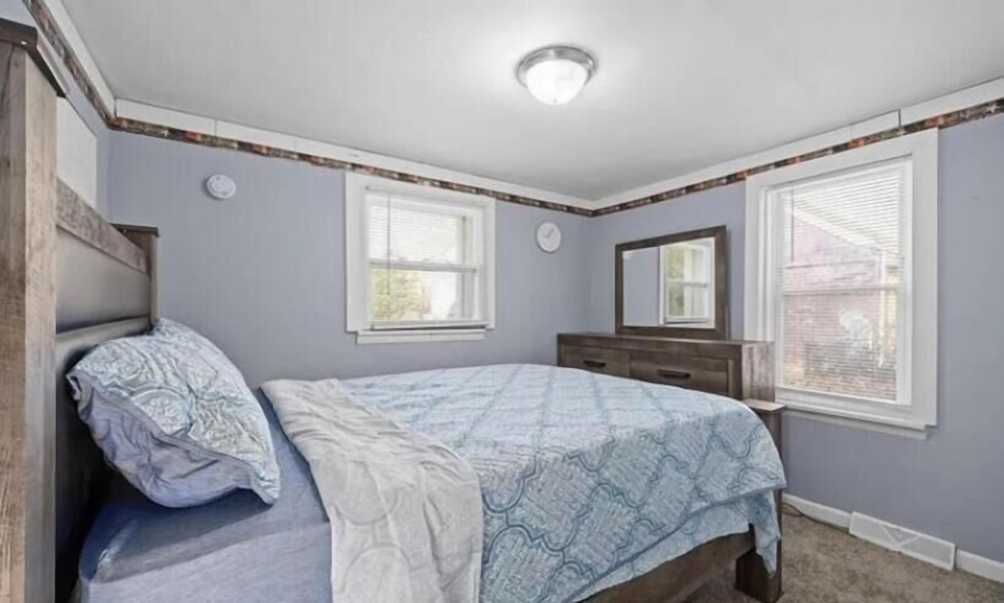 A bedroom with a bed, dresser, two windows, and a ceiling light fixture.