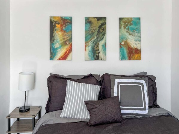 A bedroom with a bed, pillows, a bedside table with a lamp, and three abstract paintings on the wall. - 2BR Near Airport, public transit to downtown! (Boston)