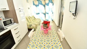 Une cuisine compacte équipée d’un four à micro-ondes, d’un four et de meubles blancs. Une table à manger, recouverte d’une nappe à motifs, est mise pour deux personnes, avec un vase de fleurs rouges en guise de décoration.