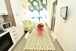 A compact kitchen with a microwave, oven, and white cabinets. A dining table with a patterned tablecloth and a vase of red flowers is set for two. - Arcobaleno Residence, Paguro Two-Room Apartment for 4 People (Chioggia)