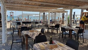 Un espace de restauration en plein air avec des tables et des chaises, une vue sur la plage et, au loin, un poste de surveillance des sauveteurs.