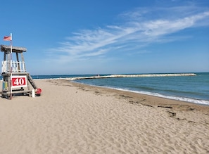 A lifeguard tower with a red flag, a clear blue sky, and a sandy beach with a rocky breakwater in the distance. - Residenz Arcobaleno, Cavalluccio Plus Three-Room Apartment for 6 People (Chioggia)