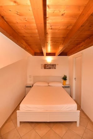 A bedroom with a wooden ceiling, a bed, a nightstand, and a wall-mounted light fixture. - Residenz Arcobaleno, Cavalluccio Plus Three-Room Apartment for 6 People (Chioggia)