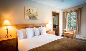 A hotel room with a bed, two bedside lamps, a wooden nightstand, a chair, a window with curtains, and a painting on the wall.