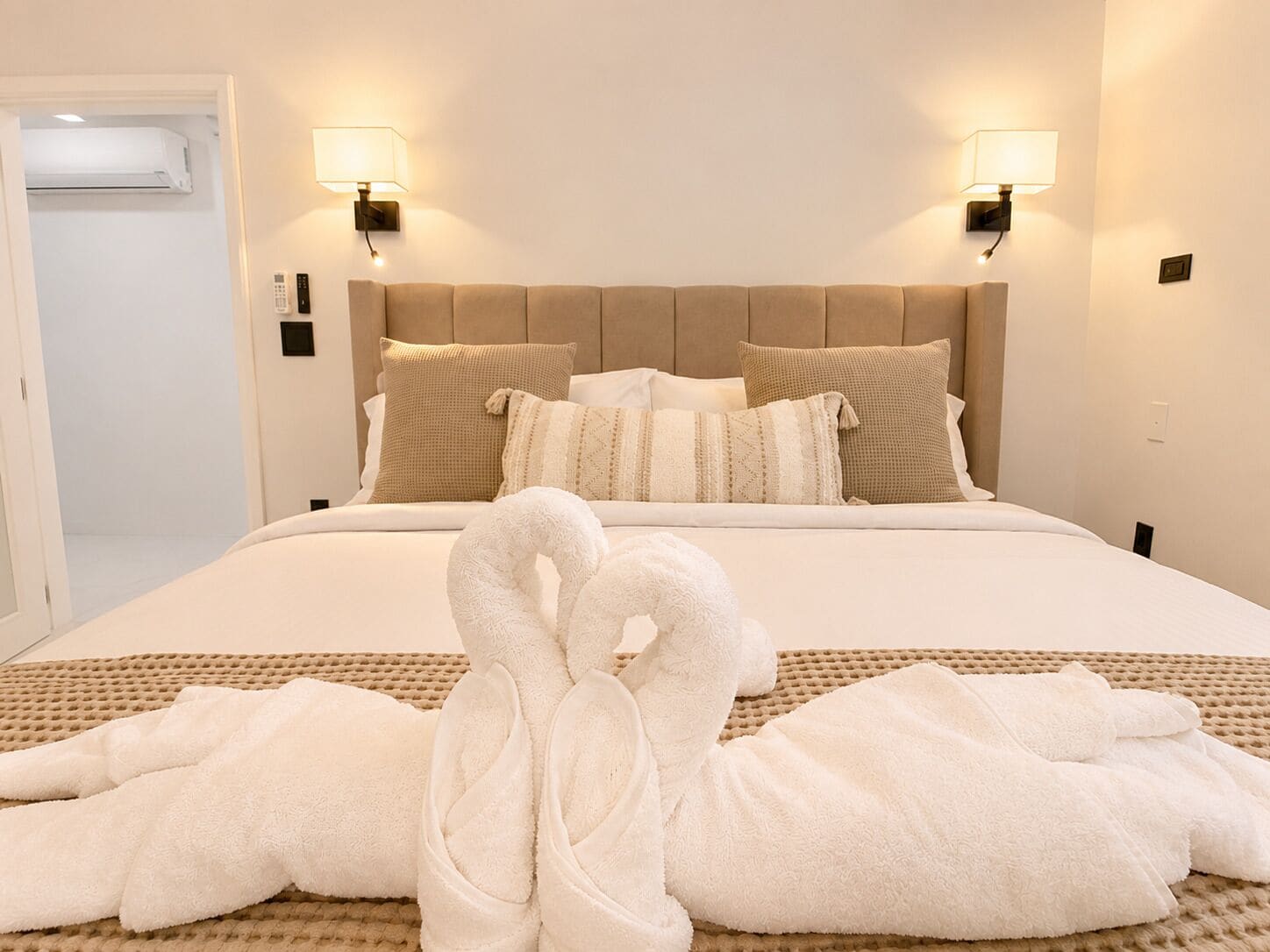 A hotel room with a bed, two wall-mounted lamps, a headboard, and a pair of towels arranged on the bed.