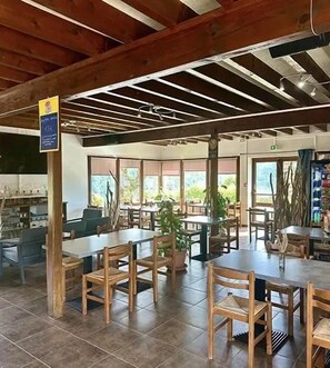 An indoor dining area with wooden tables and chairs, large windows, and a high ceiling with exposed beams.