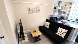Un espacio habitable compacto con sofá, mesa de centro, televisor sobre un soporte de madera, y una cocina con encimera y armarios.