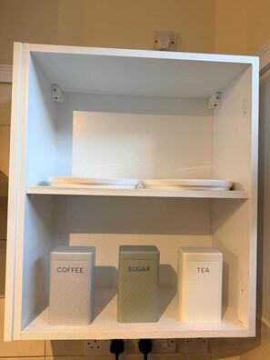 A white shelf unit with labeled containers for coffee, sugar, and tea. - Apartment Opp Leicester Train Station | Central (Leicester)