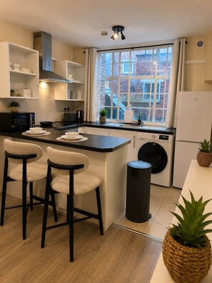 A modern kitchen with a central island, bar stools, a built-in washing machine, and a window with a view of buildings. - Apartment Opp Leicester Train Station | Central (Leicester)