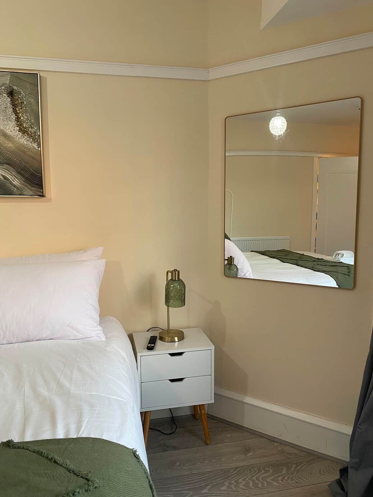 A bedroom with a bed, a bedside table with a lamp, a mirror, and a painting on the wall.