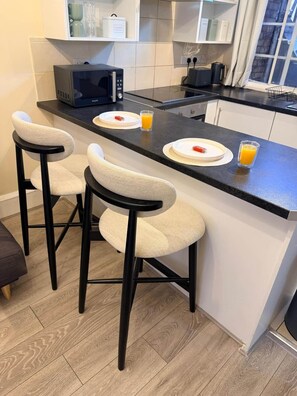 A kitchen with a black countertop, white cabinets, and a microwave. There are two bar stools with beige seats and black legs. - Apartment Opp Leicester Train Station | Central (Leicester)