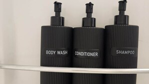 Tres botellas negras con dosificadores sobre una repisa transparente frente a una pared blanca.