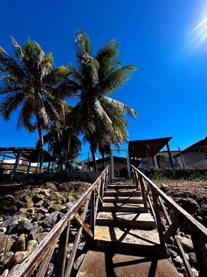 Una escalera de madera que conduce a un edificio con una verja de metal, rodeado de palmeras y rocas.