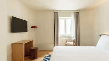 Habitación de hotel con cama, televisión montada en la pared, una mesita, una silla y ventana con cortinas.