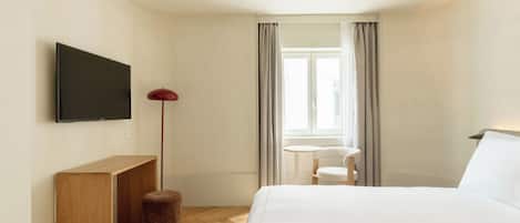 A hotel room with a bed, a TV mounted on the wall, a small table, a chair, and a window with curtains.