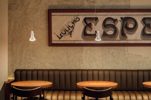 Ravintolan sisustus, jossa on puinen penkkirivi istuinalueena, kaksi pyöreää pöytää ja seinälle kiinnitetty kyltti, jossa lukee sanat »Lounges» ja »Espresso».