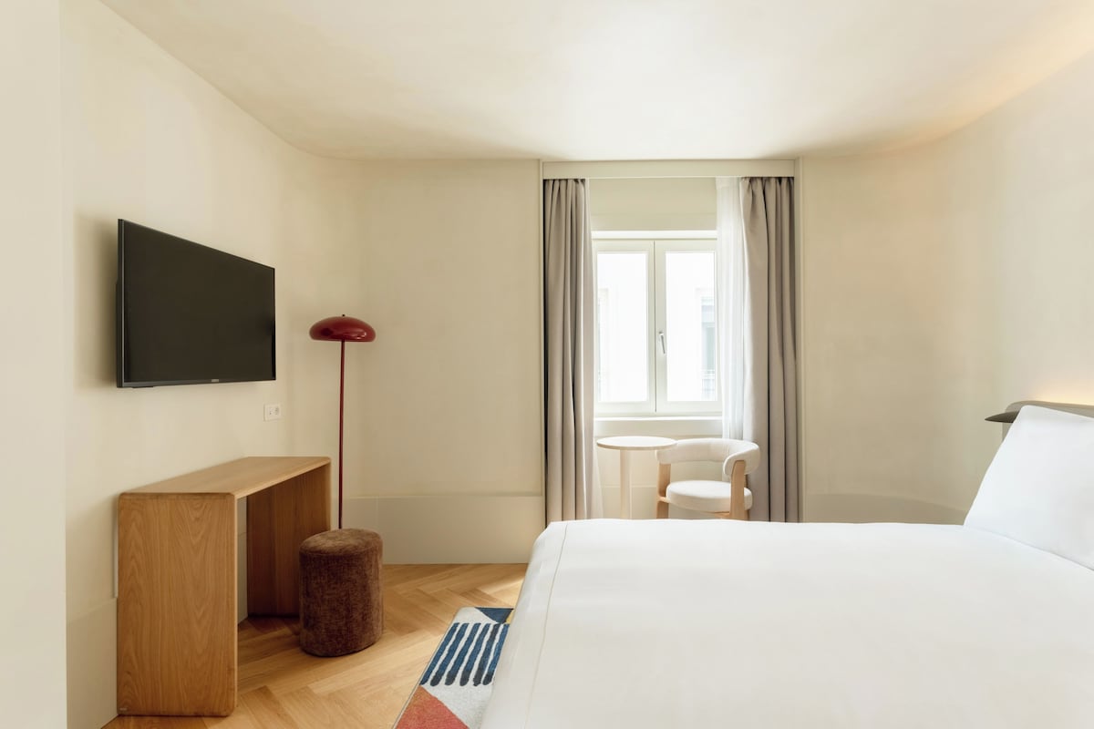Ein Hotelzimmer mit Bett, Wandfernseher, kleinem Tisch, Stuhl und einem Fenster mit Vorhängen.