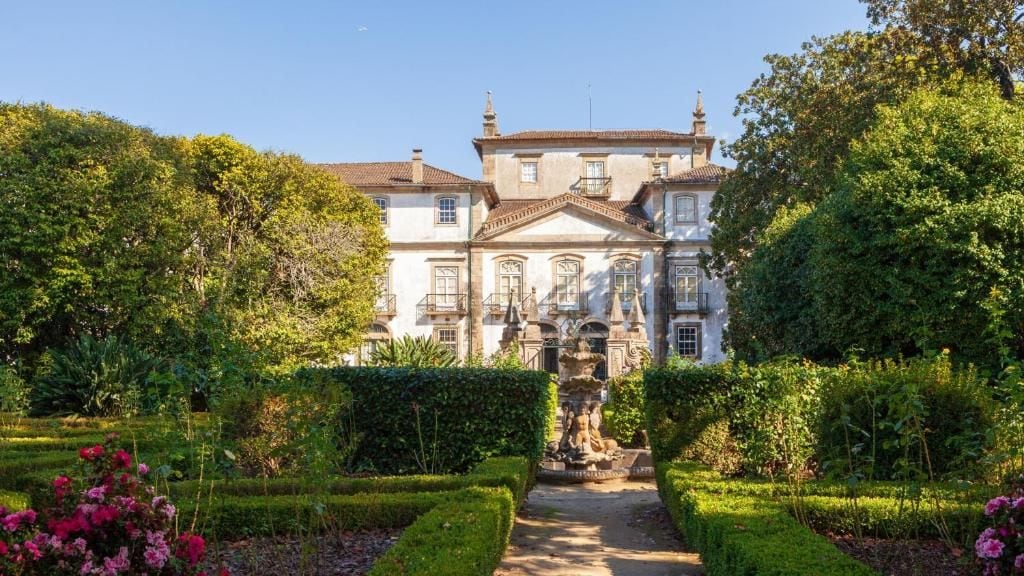 Un edificio neoclassico con una facciata simmetrica, un ingresso centrale e un giardino curato con siepi e piante fiorite.