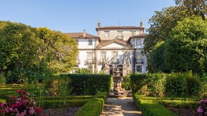 Un bâtiment de style classique, doté d’une façade symétrique, d’une entrée centrale et d’un jardin bien entretenu, agrémenté de haies et de plantes à fleurs.