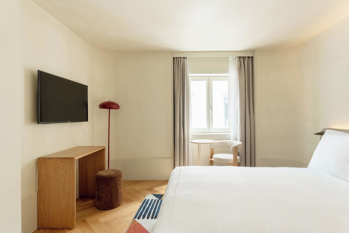 A hotel room with a bed, a TV mounted on the wall, a small table, a chair, and a window with curtains.