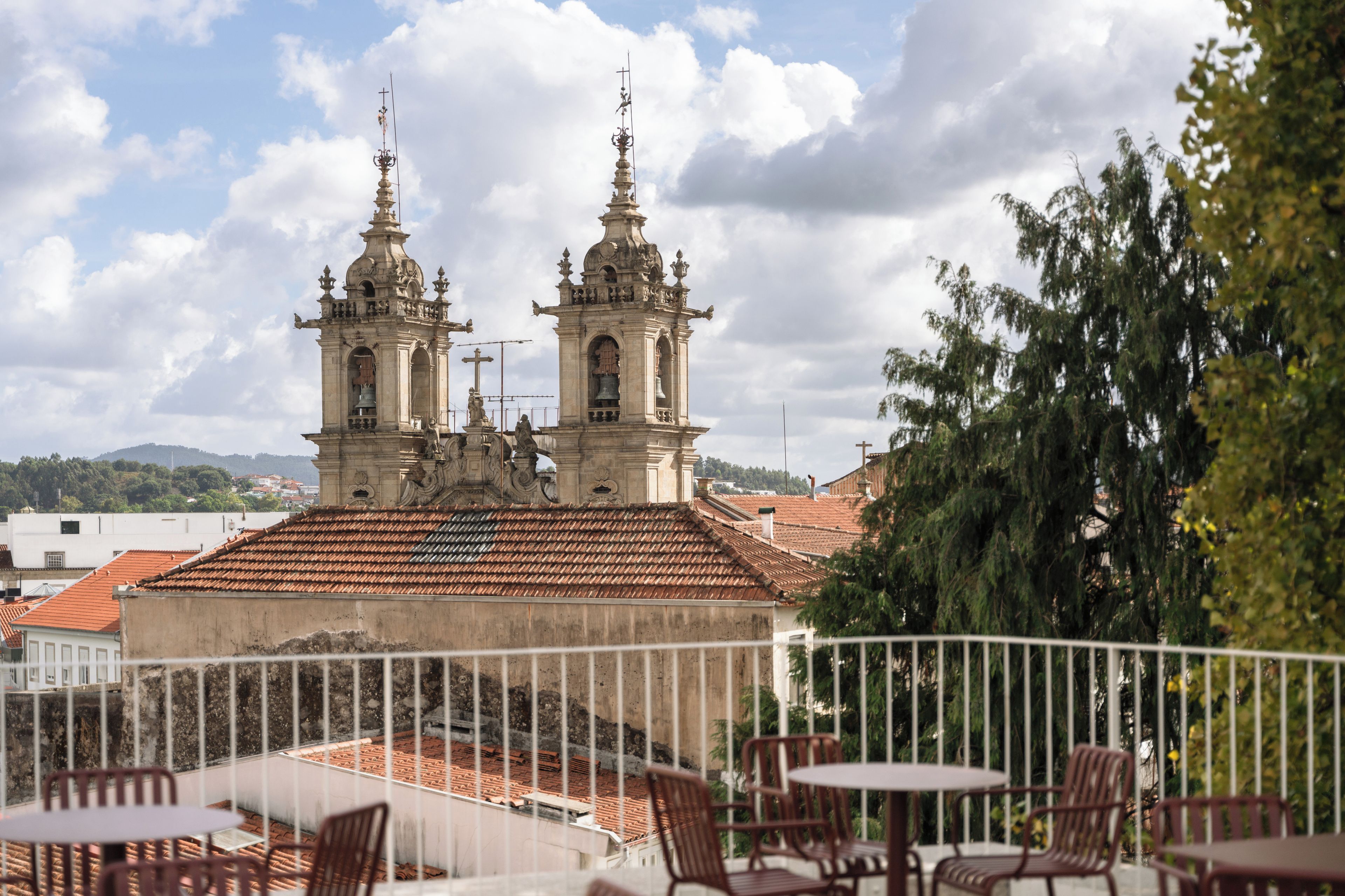 Una terraza con mesas al aire libre, con vistas a un edificio histórico con dos torres de campanas y un tejado de tejas rojas.